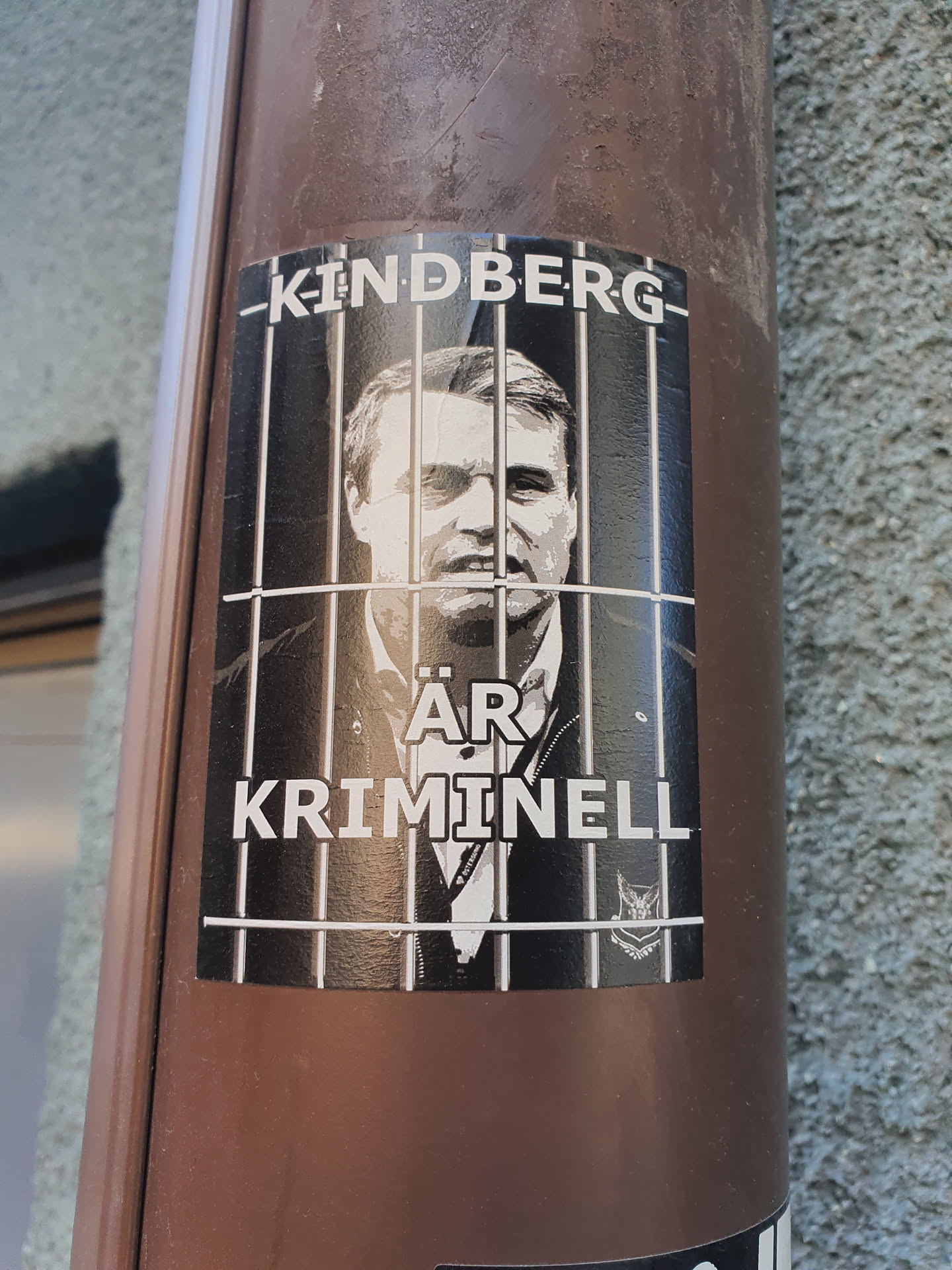 Black and white sticker depicting a man behind bars with the text "Kindberg är kriminell" (Kindberg is a criminal) in Swedish. The sticker has a logo at the bottom right corner resembling a stylized bird of prey.