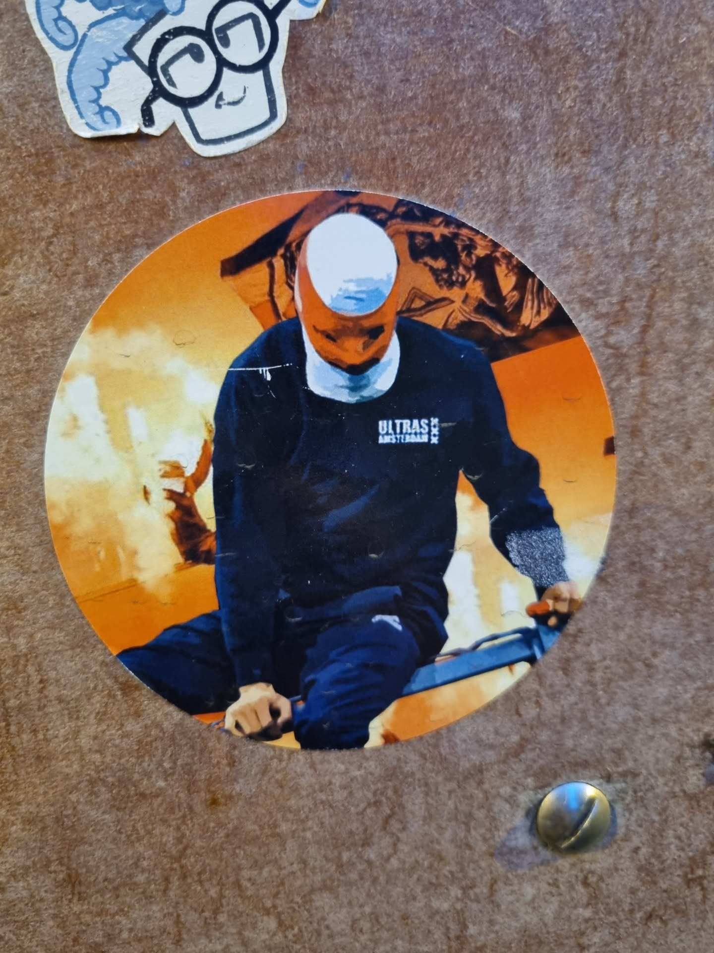 Round sticker depicting a stylized image of a person riding a bike, possibly BMX or similar, against a backdrop of intense orange and yellow flames. The person is wearing dark clothing with the word 'ULTRAS' and 'AMSTERDAM' visible on their shirt. The overall style is bold and graphic.