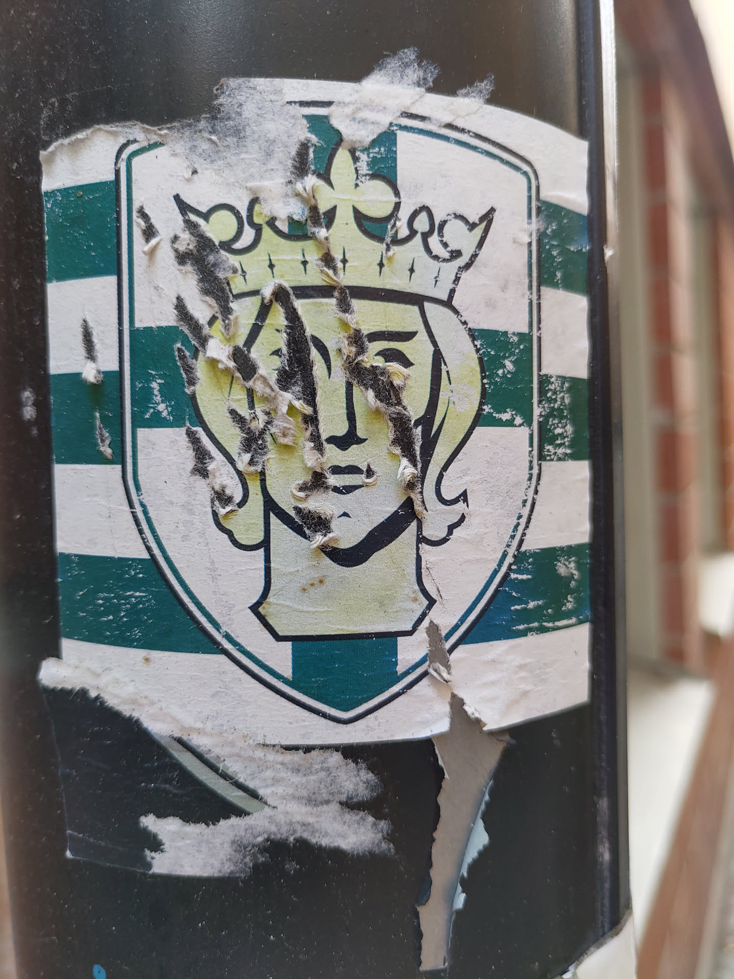 This sticker shows a worn-out emblem with a woman wearing a crown inside a shield-shaped design.  The colors are mainly green and white. The sticker is significantly damaged and peeling.