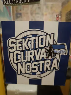 A sticker featuring the logo of Sektion Curva Nostra, a Hertha BSC fan group. The design includes the group's name, a bear emblem, the club's abbreviation (Hertha BSC), and the year 1892.  The background is blue and white vertical stripes.