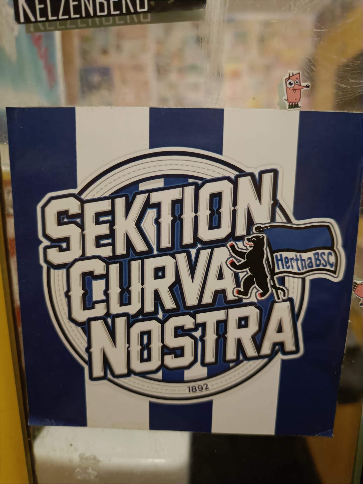 A sticker featuring the logo of Sektion Curva Nostra, a Hertha BSC fan group. The design includes the group's name, a bear emblem, the club's abbreviation (Hertha BSC), and the year 1892.  The background is blue and white vertical stripes.
