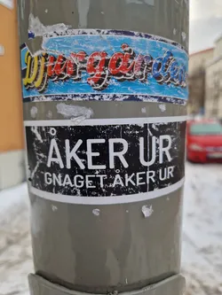 A black and white sticker with the text ÅKER UR and Gnag et ÅKER UR underneath.  It's affixed to a grey pole, partially obscuring another sticker with illegible text above it.