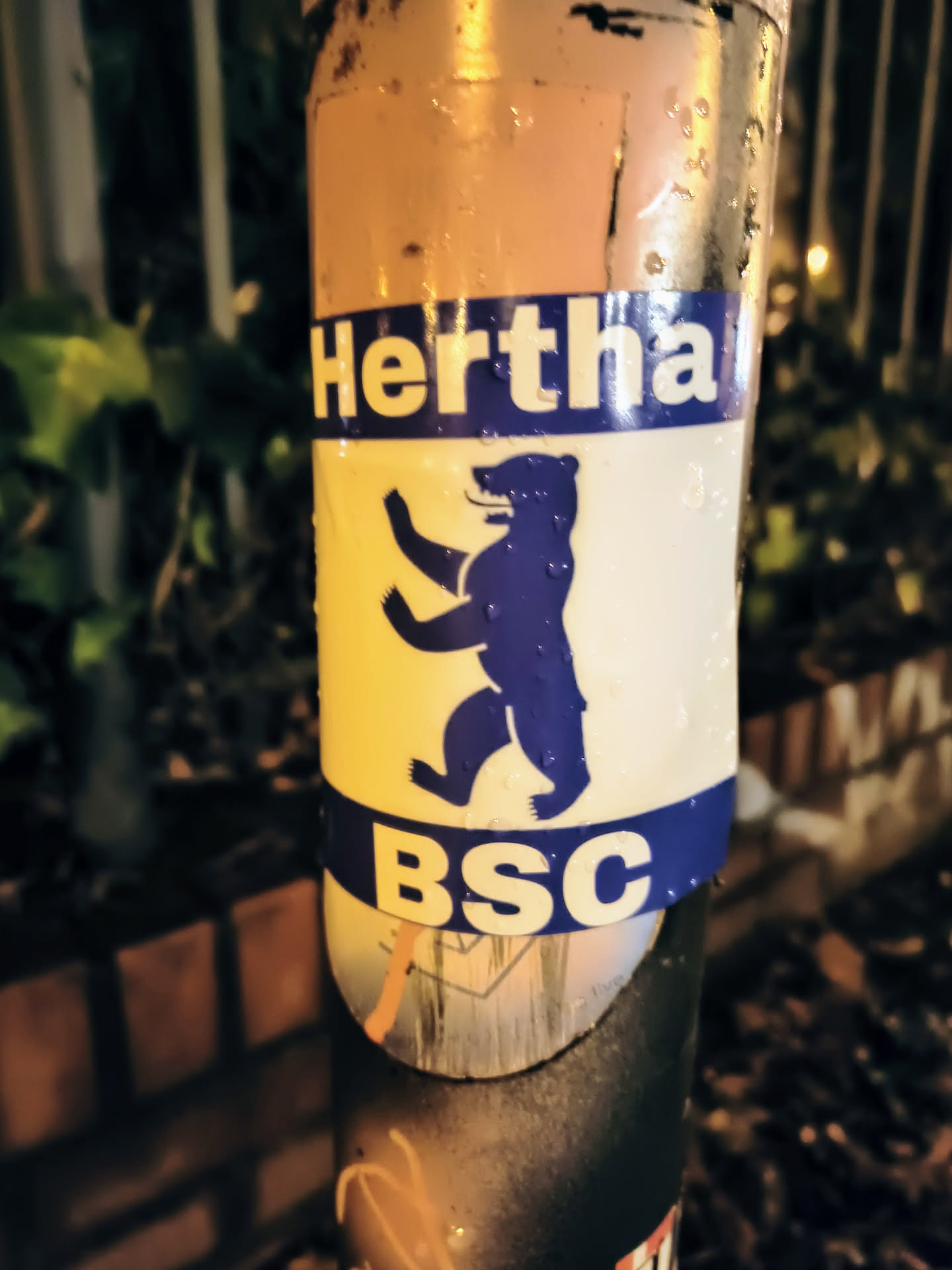 A sticker featuring the Hertha BSC logo, a blue bear on a yellow background, with 'Hertha' and 'BSC' written in blue lettering. The sticker is affixed to a weathered pole.