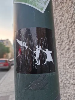 A black and white sticker featuring silhouettes of figures, possibly engaged in a struggle or dance. The style is bold and graphic. The sticker is affixed to a pole outdoors.