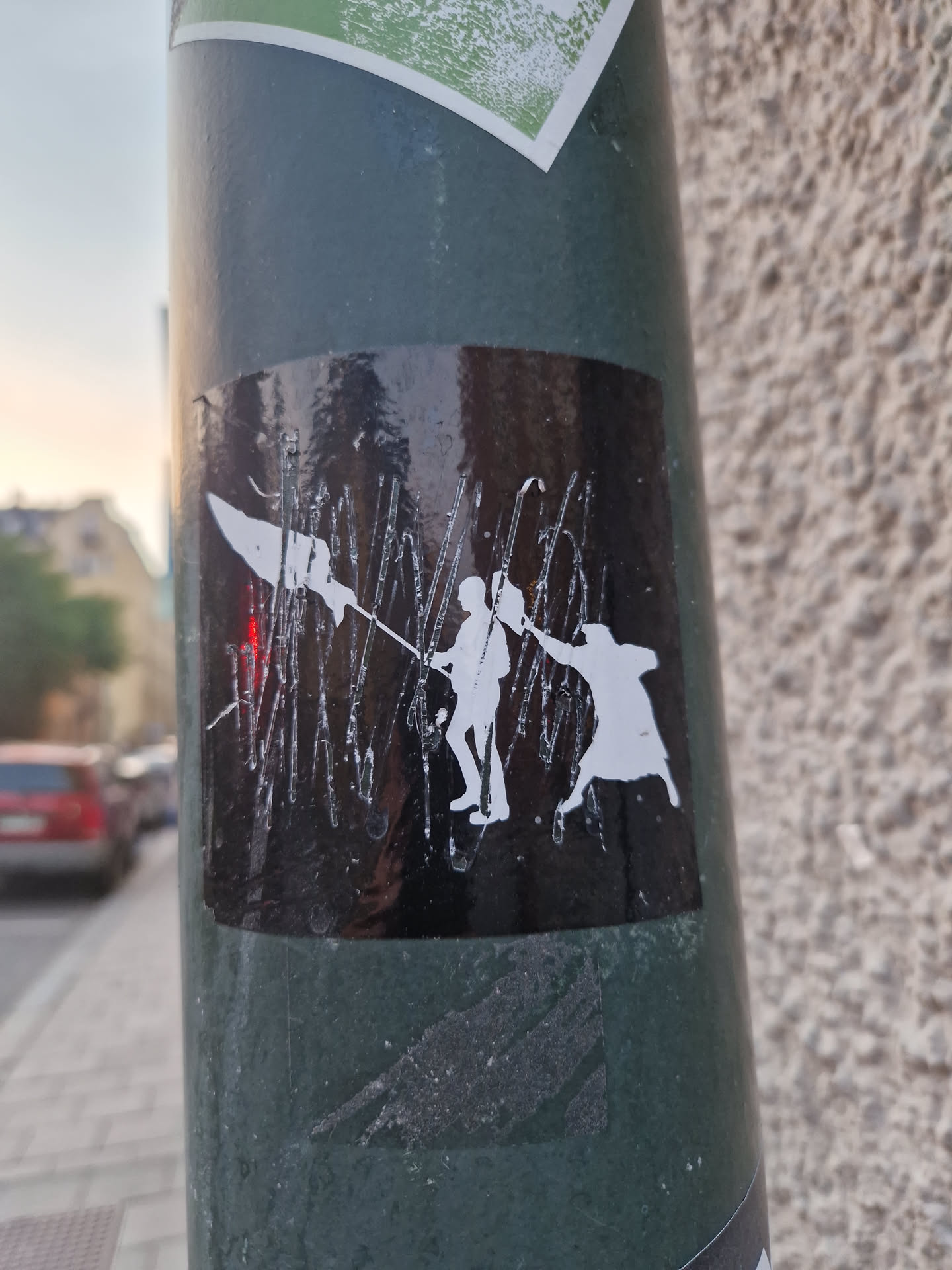 A black and white sticker featuring silhouettes of figures, possibly engaged in a struggle or dance. The style is bold and graphic. The sticker is affixed to a pole outdoors.