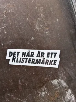 A rectangular sticker with a white background and bold black text. The text is split into two lines, with the top line 'DET HÄR ÄR ETT' slightly offset to the left above the bottom line 'KLISTERMÄRKE'. The sticker has clean-cut edges and is applied to a dark, scratched, and somewhat dusty brown surface.