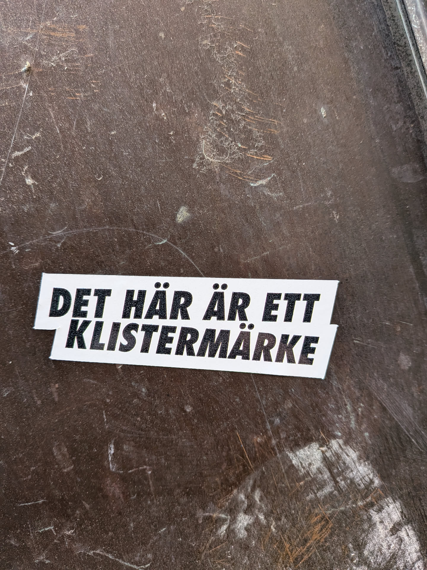 A rectangular sticker with a white background and bold black text. The text is split into two lines, with the top line 'DET HÄR ÄR ETT' slightly offset to the left above the bottom line 'KLISTERMÄRKE'. The sticker has clean-cut edges and is applied to a dark, scratched, and somewhat dusty brown surface.