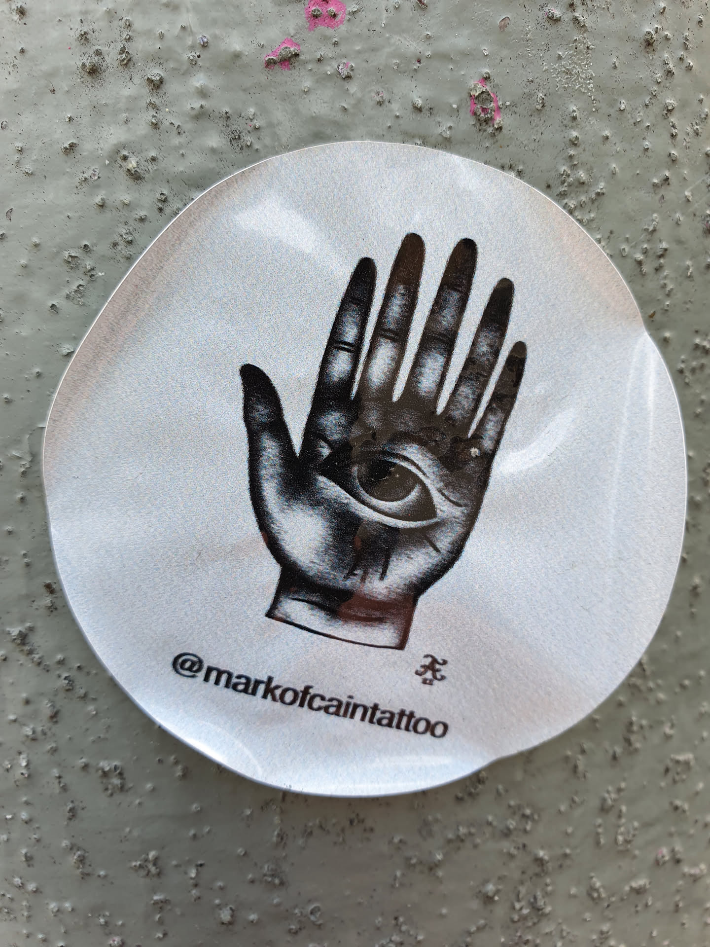 A round sticker featuring a black and white illustration of a hand with an eye in the palm. The sticker has a slightly reflective surface. The Instagram handle @markofcaintattoo is printed at the bottom of the sticker.