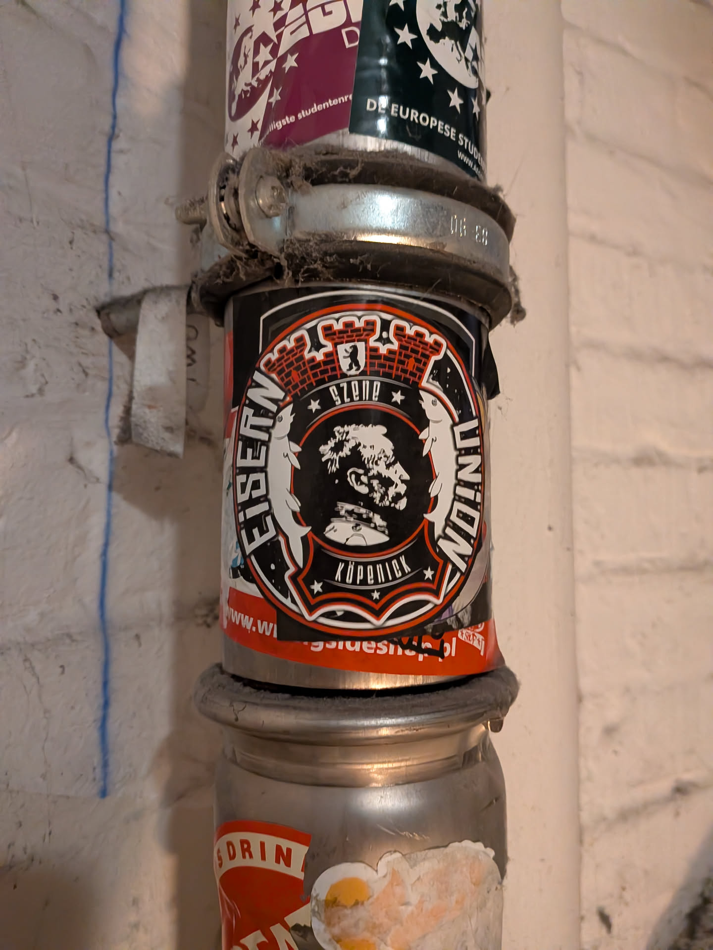 A circular sticker representing fans of 1. FC Union Berlin. The design is styled like an emblem or crest, featuring a red brick mural crown at the top with a small Berlin bear shield. Flanking the sides are two heraldic fish from the Köpenick coat of arms. The text 'EISERN' and 'UNION' curves along the outer ring. The inner circle displays a silhouette bust of the Hauptmann von Köpenick (Captain of Köpenick) above a banner reading 'KÖPENICK', with 'SZENE' centered above between two stars.