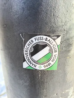 A sticker with the Hannover 96 football club logo is affixed to a dark gray metal post. The logo features the club name encircling a shield with black and green stripes, along with the founding year of 1896.
