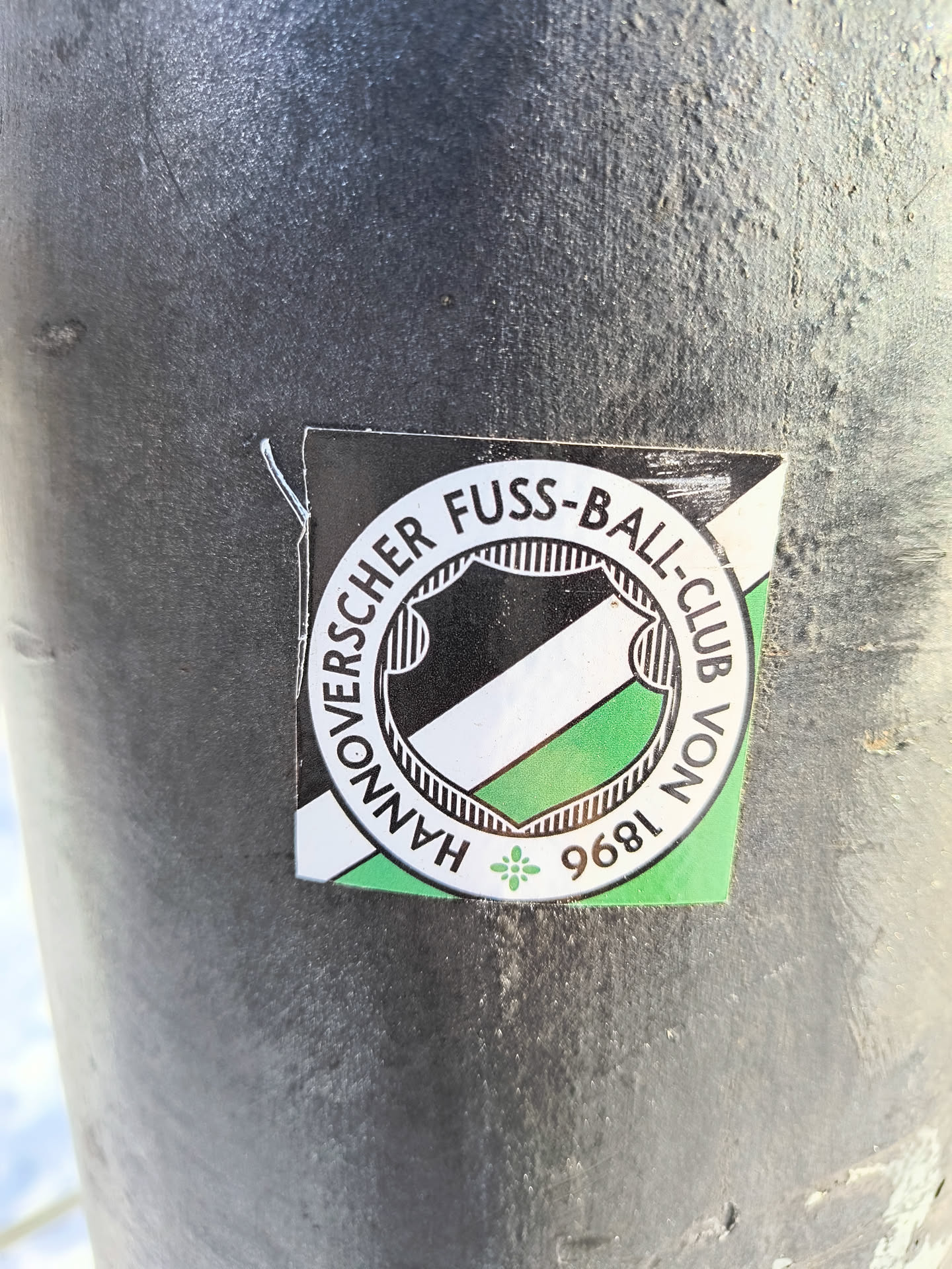 A sticker with the Hannover 96 football club logo is affixed to a dark gray metal post. The logo features the club name encircling a shield with black and green stripes, along with the founding year of 1896.