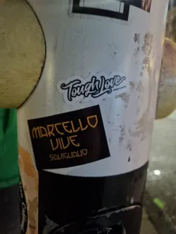 Street sticker A black and gold sticker with the text "MARCELLO VIVE" stacked over "SAUGUADLO" on a black background.  Above it is a white sticker with the text "Tough Love" written in a stylized script font.