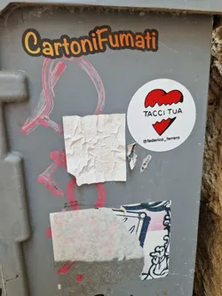 A white round sticker with a red broken heart and the text "TACCI TUA". The sticker is attached to a gray surface with other graffiti and stickers around it.  There's also a small instagram handle visible below the main text.