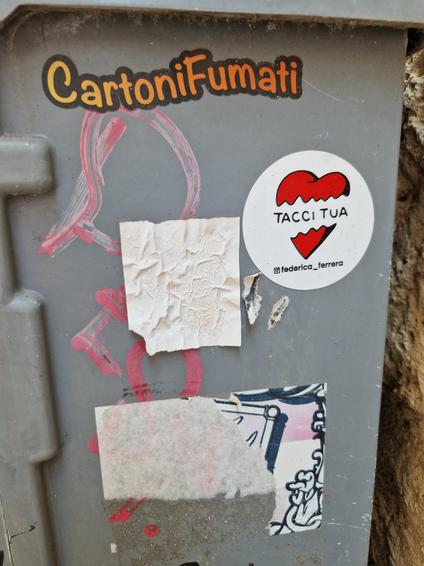 A white round sticker with a red broken heart and the text "TACCI TUA". The sticker is attached to a gray surface with other graffiti and stickers around it.  There's also a small instagram handle visible below the main text.