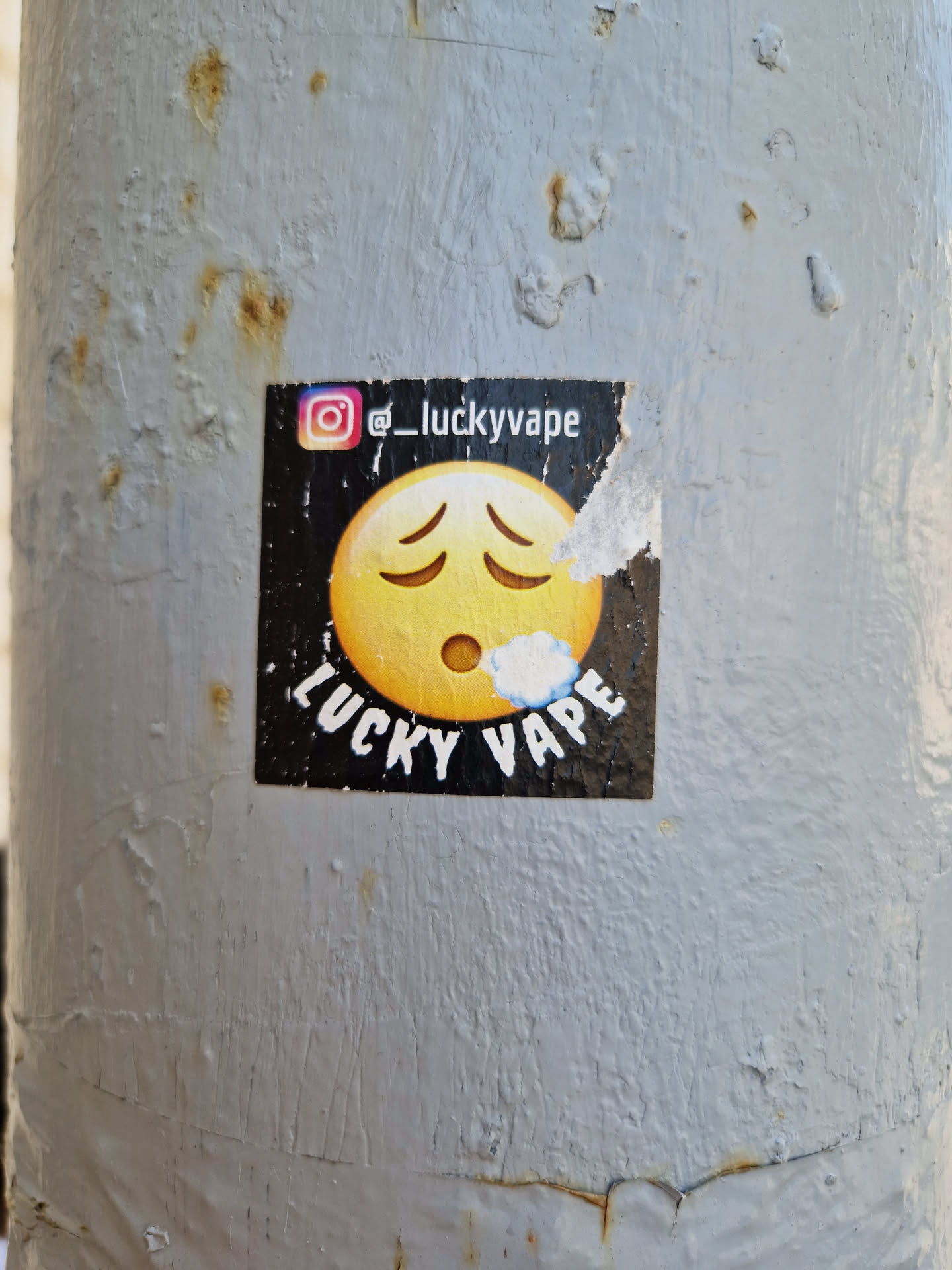 A sticker with a distressed emoticon and the text "Lucky Vape" and Instagram handle @luckyvape. The sticker is placed on a weathered metal pole.