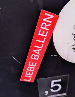 A rectangular red sticker featuring the German phrase "LIEBE BALLERN" in white capital letters. The sticker is placed on a dark, possibly black, surface alongside other stickers. The sticker has a minimalist design with a bold contrast between the red background and the white text.