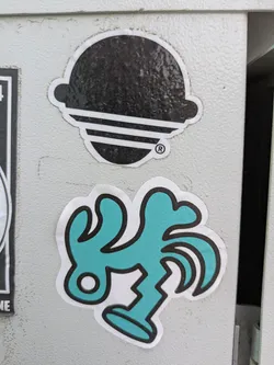 The image features a teal-colored sticker with a quirky, abstract design. It has a white border and is adhered to a gray metal surface. The design appears to be a stylized figure or creature with irregular shapes and lines, giving it a playful and unconventional aesthetic. Above it is another sticker which is mostly black.