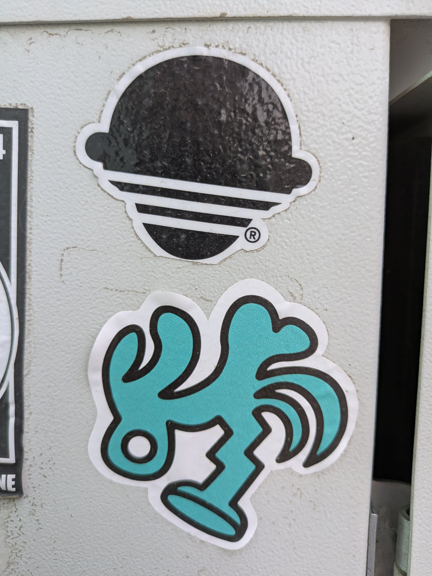 The image features a teal-colored sticker with a quirky, abstract design. It has a white border and is adhered to a gray metal surface. The design appears to be a stylized figure or creature with irregular shapes and lines, giving it a playful and unconventional aesthetic. Above it is another sticker which is mostly black.