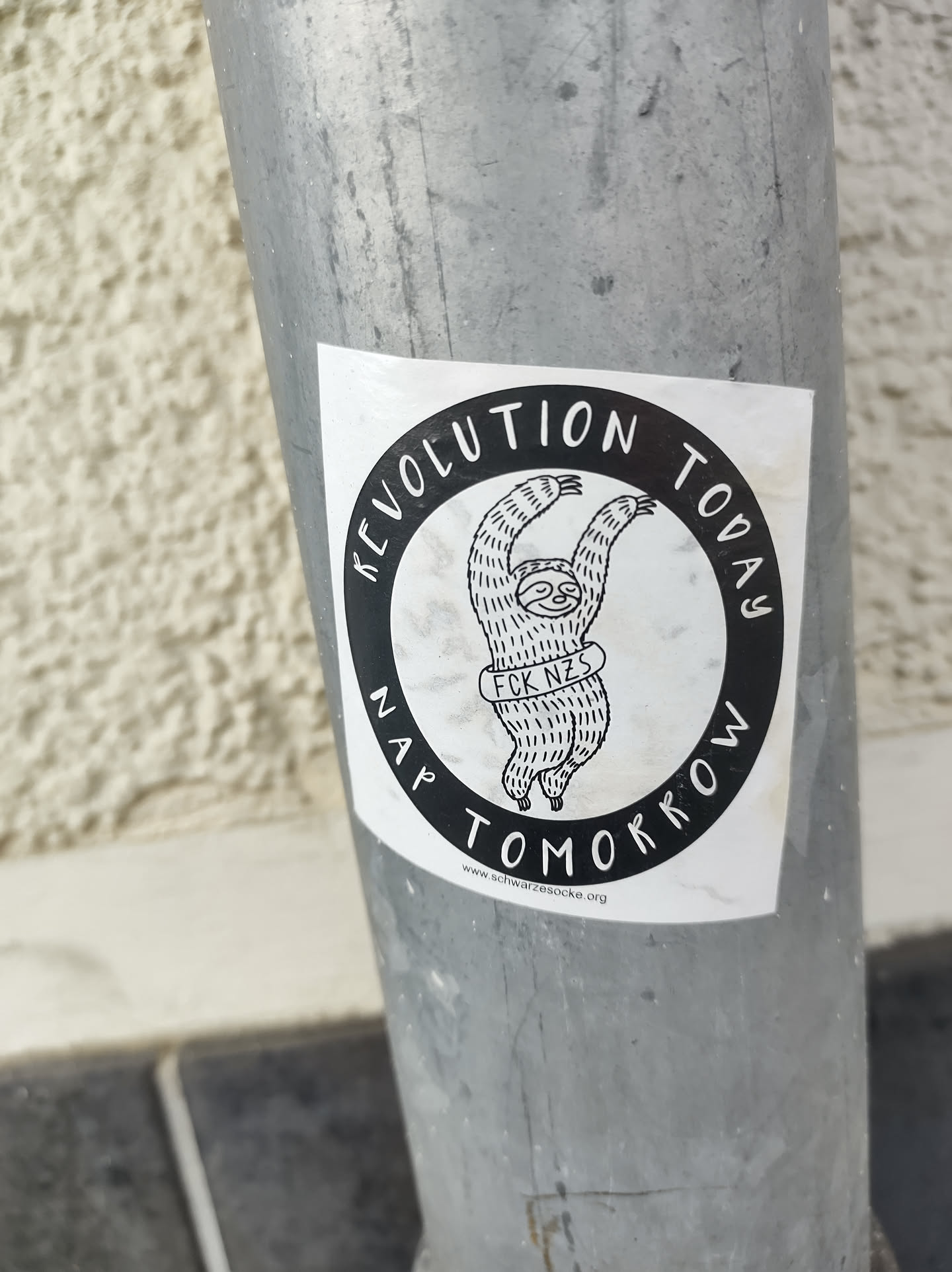 A black and white sticker featuring a sloth with the text "Revolution Today, Nap Tomorrow" and "FCK NZS" written on it. The sticker is attached to a metal pole.