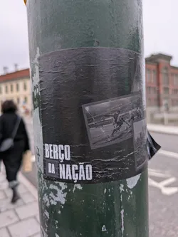A black and white image of a historical sporting event, possibly a horse race, is featured on a worn black sticker. The text "BERÇO DA NAÇÃO" is prominently displayed. The sticker appears to be affixed to a weathered green pole.