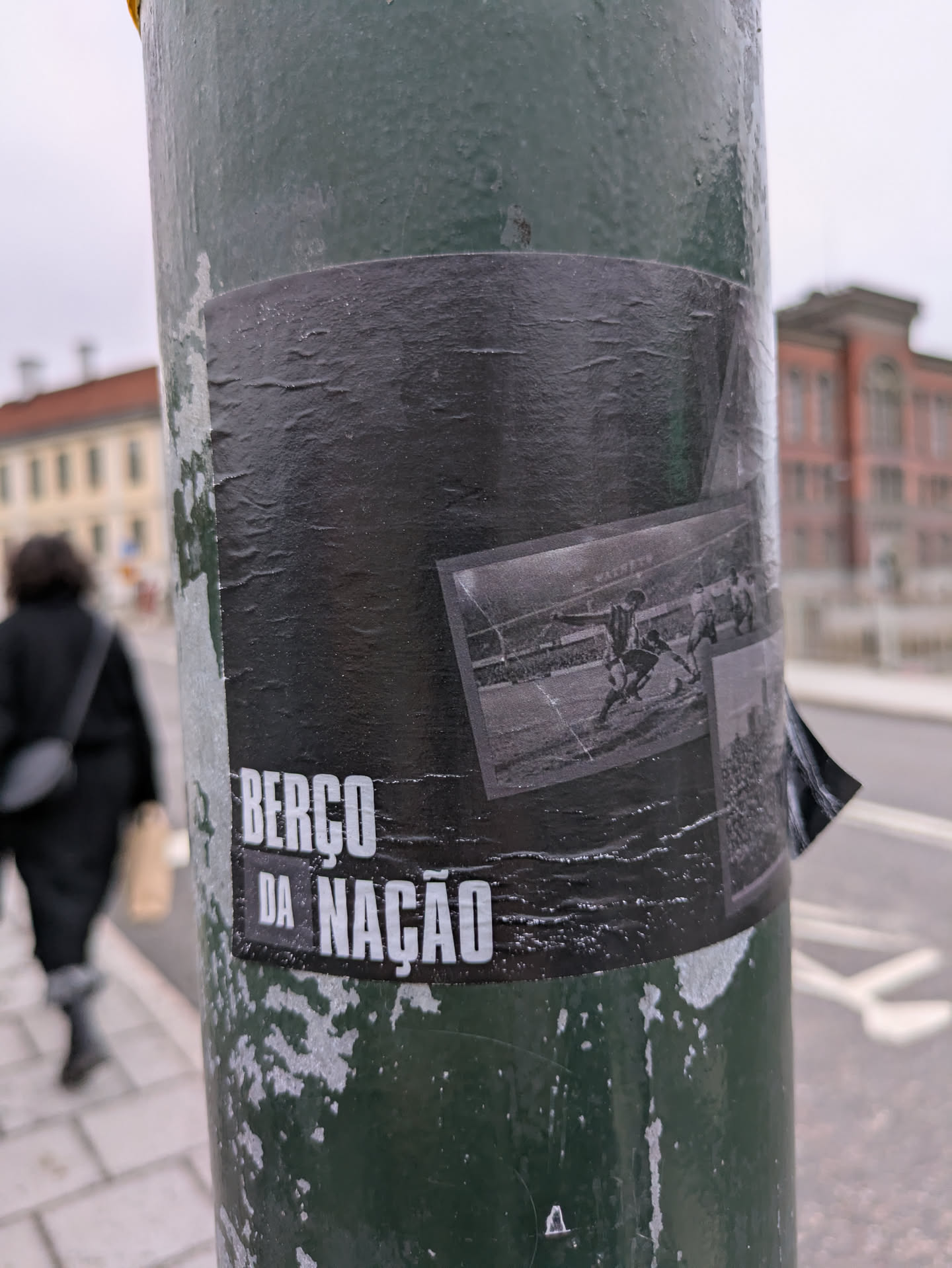 A black and white image of a historical sporting event, possibly a horse race, is featured on a worn black sticker. The text "BERÇO DA NAÇÃO" is prominently displayed. The sticker appears to be affixed to a weathered green pole.