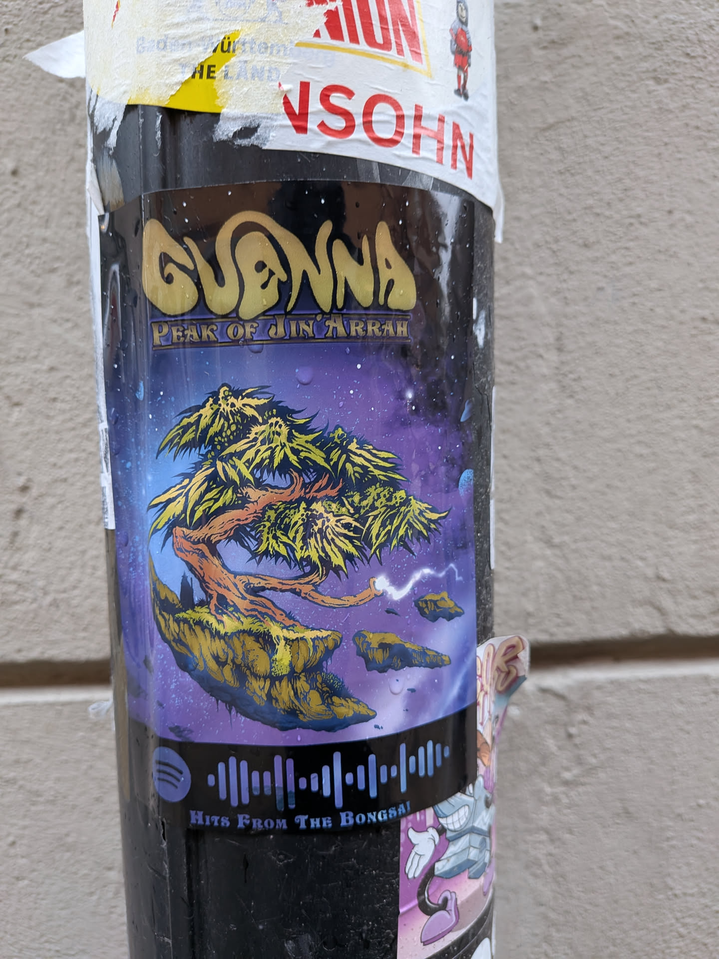 The sticker features a vibrant, fantastical scene set against a deep purple and blue cosmic background dotted with numerous white stars and nebulae. At its center is a gnarled, ancient-looking tree with bright yellow-green, spiky foliage, rooted on a large, textured floating island of rock and earth. Below the main island, smaller rock fragments drift, with a streak of white energy or lightning emanating from one of them. Above the main artwork, the band name "GUANNA" is written in a stylized, golden, bubbly font, with "PEAK OF JIN ARRAH" beneath it in a smaller, more angular script. At the bottom, a light blue Spotify audio wave code is prominently displayed, accompanied by the text "HITS FROM THE BONGSAI" in a similar font. The sticker appears glossy and has visible water droplets, suggesting it's exposed to the elements on a pole.