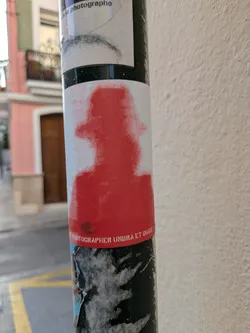 A red silhouette sticker featuring a figure with a hat, adhered to a pole. The sticker has the text 'PHOTOGRAPHER UMBRA ET IMAGO' on it.