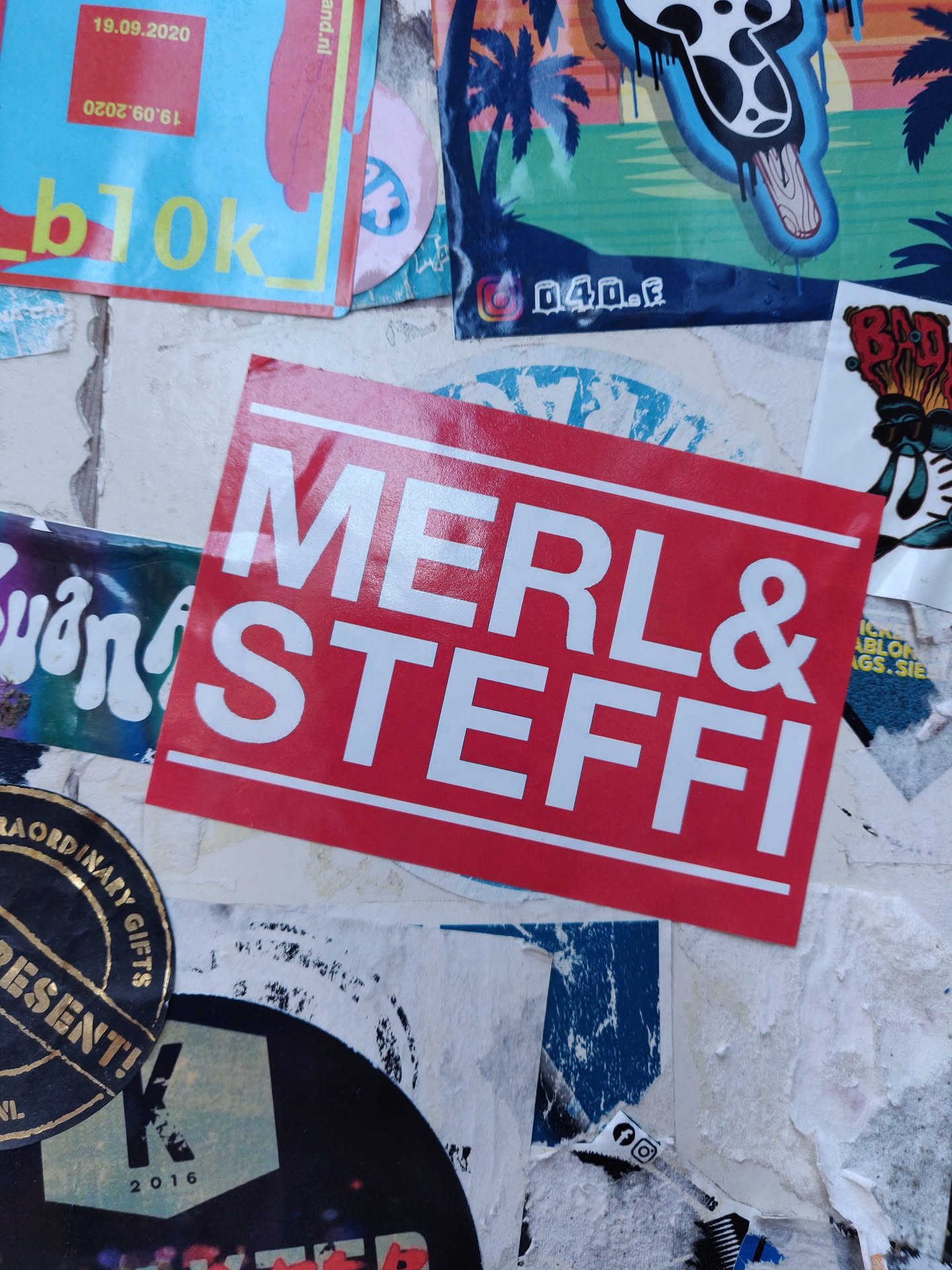 A red square sticker with white text. The text says "MERL & STEFFI" in a bold, sans-serif font. The sticker has a simple, clean design.
