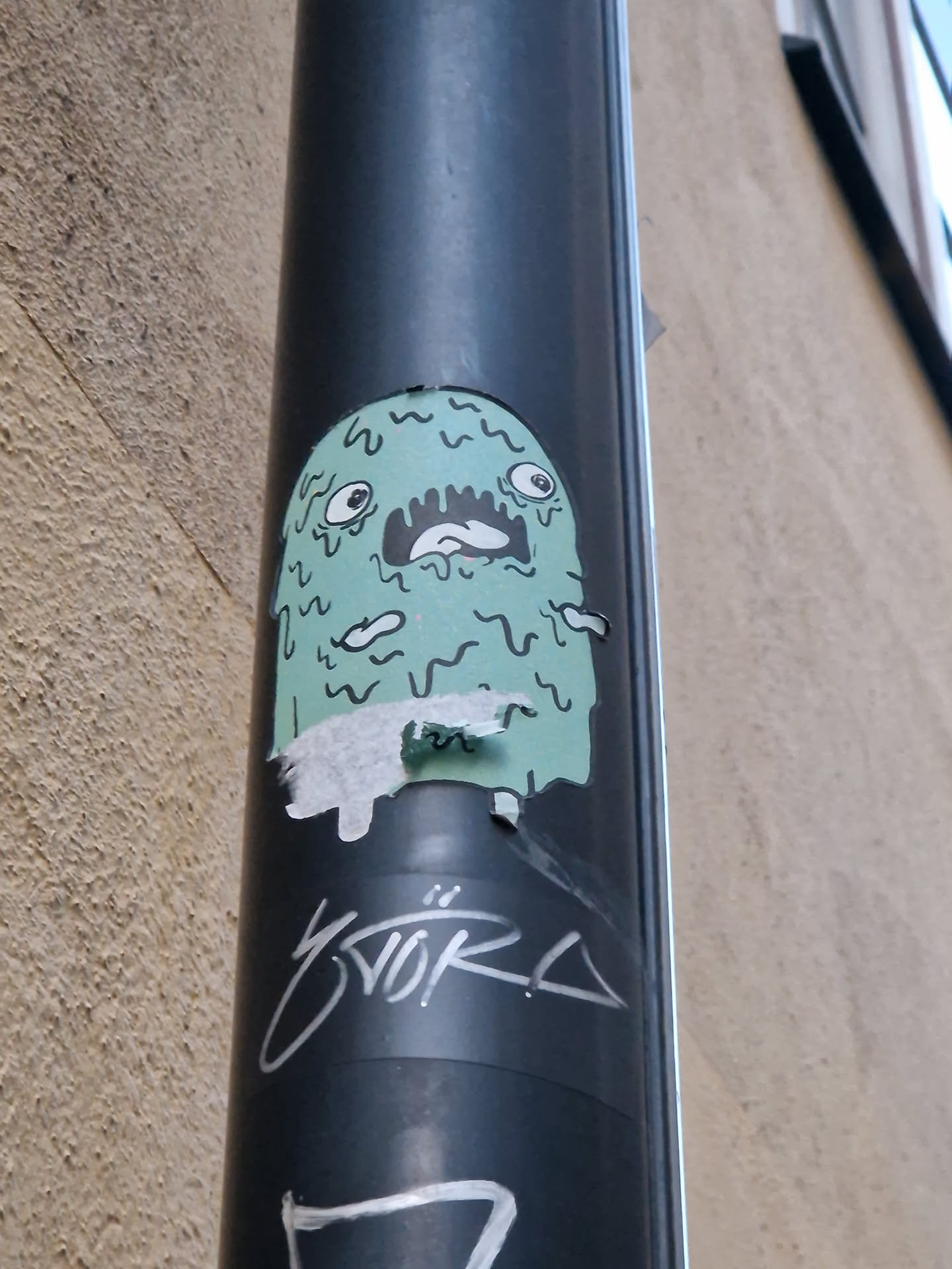 A light teal monster sticker with dripping features is affixed to a dark pole. The monster has a surprised expression. There is some graffiti below the sticker.