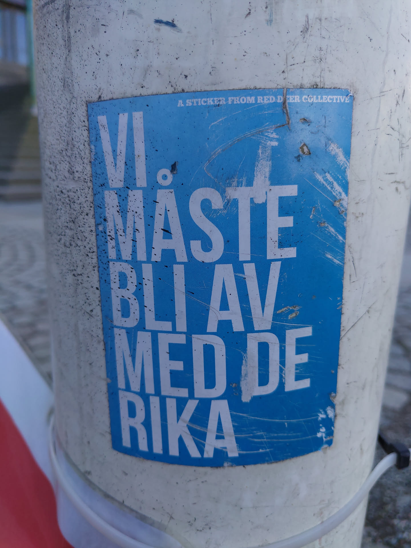 A blue sticker with white text on it. The text says "Vi måste bli av med de rika", which translates to "We must get rid of the rich". The sticker is placed on a pole. At the top it says "A sticker from Redder Collective".