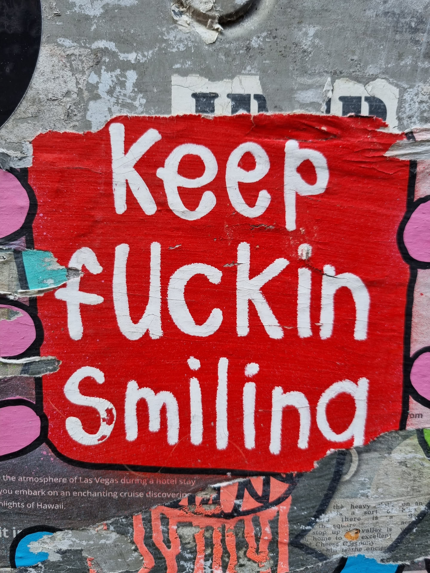 A red sticker with white hand-painted text. The sticker is slightly damaged and torn at the edges. It's affixed to a wall that shows other layers of stickers and posters underneath.