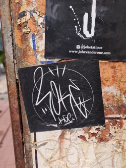 A black sticker with white graffiti art inside a circle. The signature KBC is visible at the bottom of the design. The sticker is on a rusty surface with other stickers around it. There is another sticker above with a social media handle and website.
