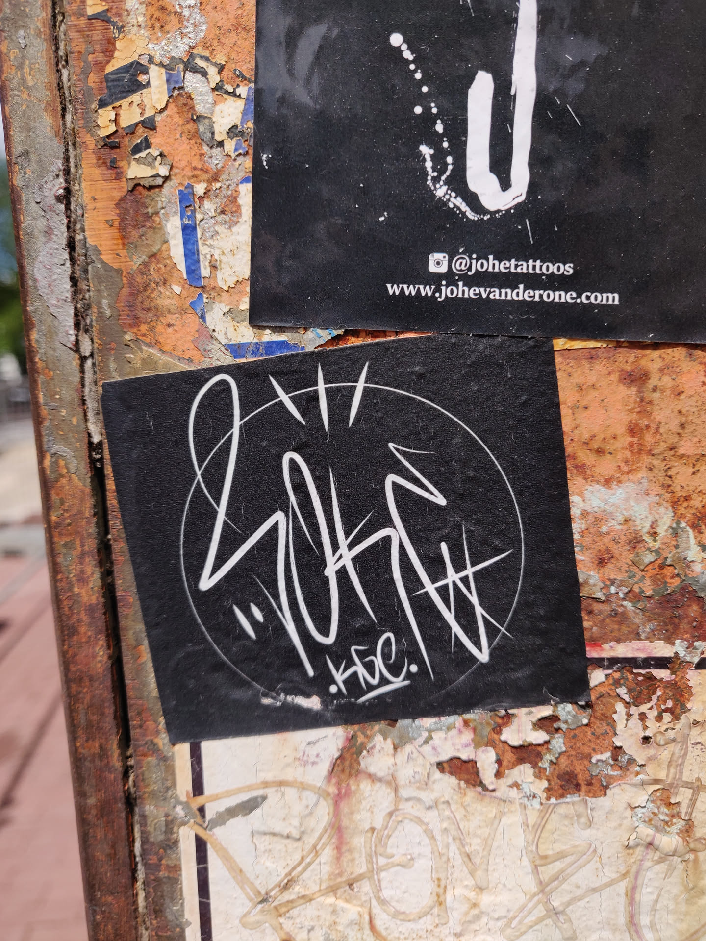 A black sticker with white graffiti art inside a circle. The signature KBC is visible at the bottom of the design. The sticker is on a rusty surface with other stickers around it. There is another sticker above with a social media handle and website.