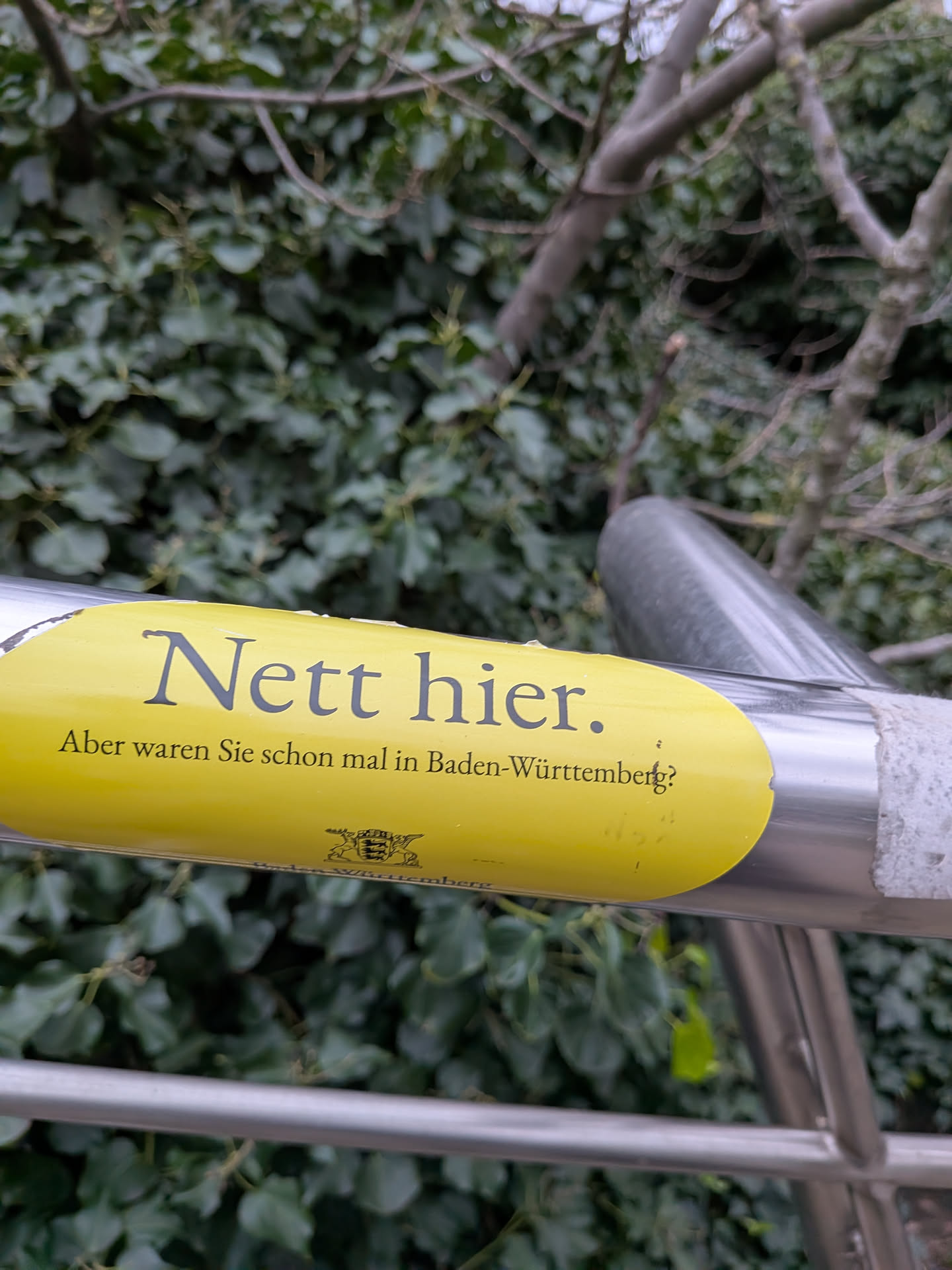 The sticker is an oval-shaped, bright yellow decal affixed to a silver metal pole. It features two lines of text in a dark grey or black serif font. The top line reads 'Nett hier.' (Nice here.) and the second line, slightly smaller, reads 'Aber waren Sie schon mal in Baden-Württemberg?' (But have you ever been to Baden-Württemberg?). Below the text, a small, intricate black crest, resembling the coat of arms of Baden-Württemberg, is visible. The sticker shows minor signs of wear, with some dirt and slight peeling at the edges, suggesting it's a physical sticker.