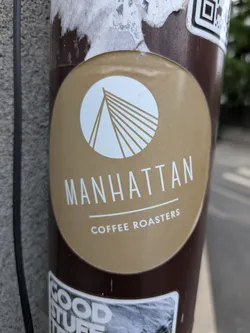 A round sticker for Manhattan Coffee Roasters, featuring a white bridge design inside a circle above the text "MANHATTAN" and "COFFEE ROASTERS" on a brown background. The sticker is affixed to a pole, and there's a QR code sticker above it, along with another sticker below.