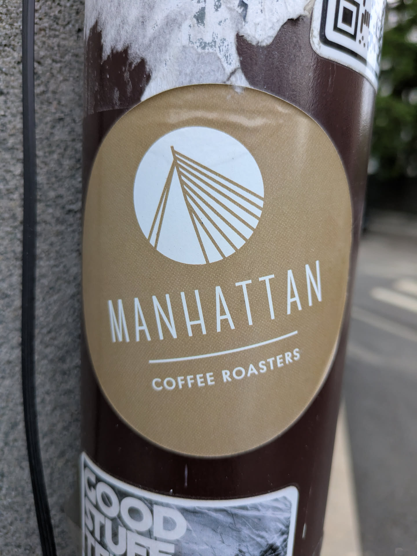 A round sticker for Manhattan Coffee Roasters, featuring a white bridge design inside a circle above the text "MANHATTAN" and "COFFEE ROASTERS" on a brown background. The sticker is affixed to a pole, and there's a QR code sticker above it, along with another sticker below.