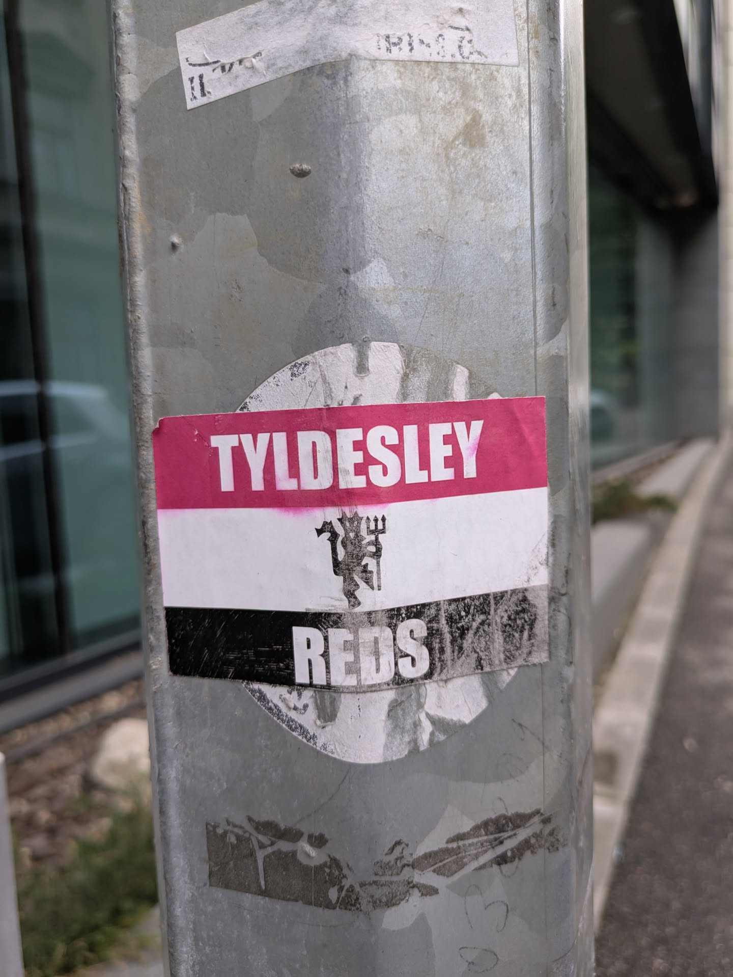 A sticker found on a metal pole. The sticker is a tricolor with the words 'TYLDESLEY' on a pink background at the top, a white middle section with a stylized devil logo and 'REDS' in white letters on a black background at the bottom.
