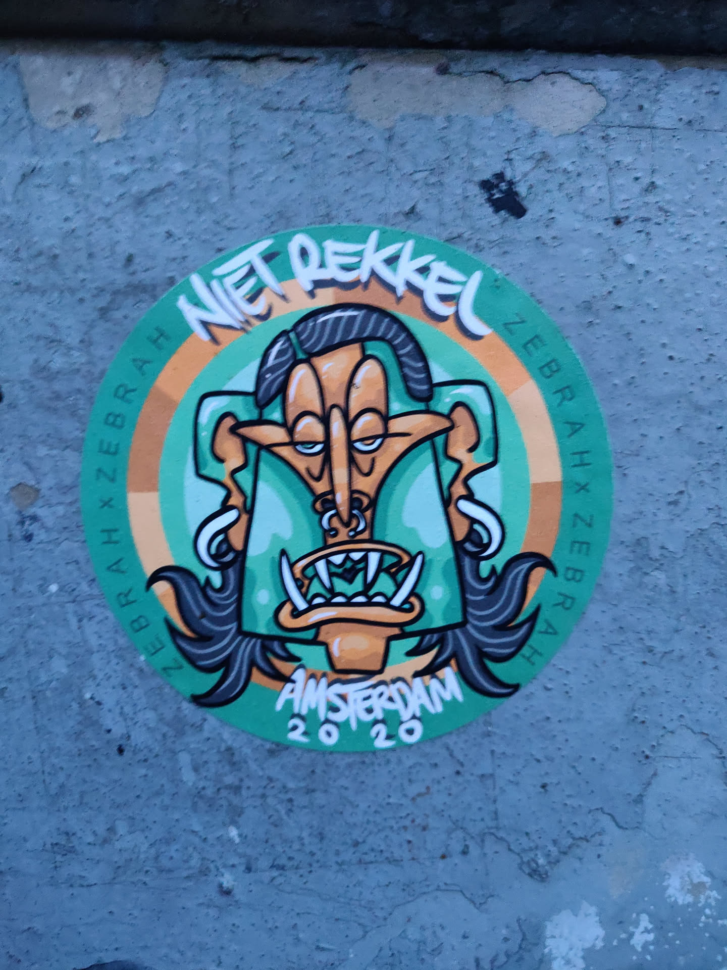 Round sticker featuring a stylized cartoon character with exaggerated features. The character has long black hair, a large nose, and prominent teeth. The text Niet Pekkel is written at the top, and Amsterdam 2020 at the bottom. The background is teal with orange accents.  The text Zebra x Zebra is repeated around the edge.