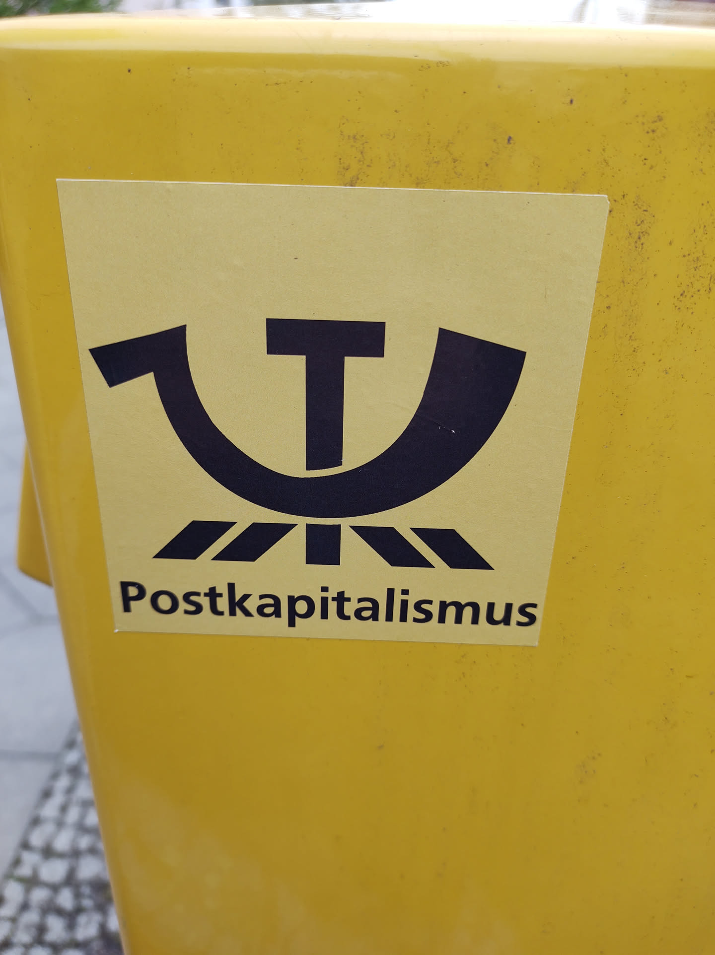 A yellow sticker with a black symbol and the word "Postkapitalismus" (post-capitalism) in German. The symbol seems to be a combination of an upside-down 'U' and a 'T' possibly representing a concept related to post-capitalist ideologies.