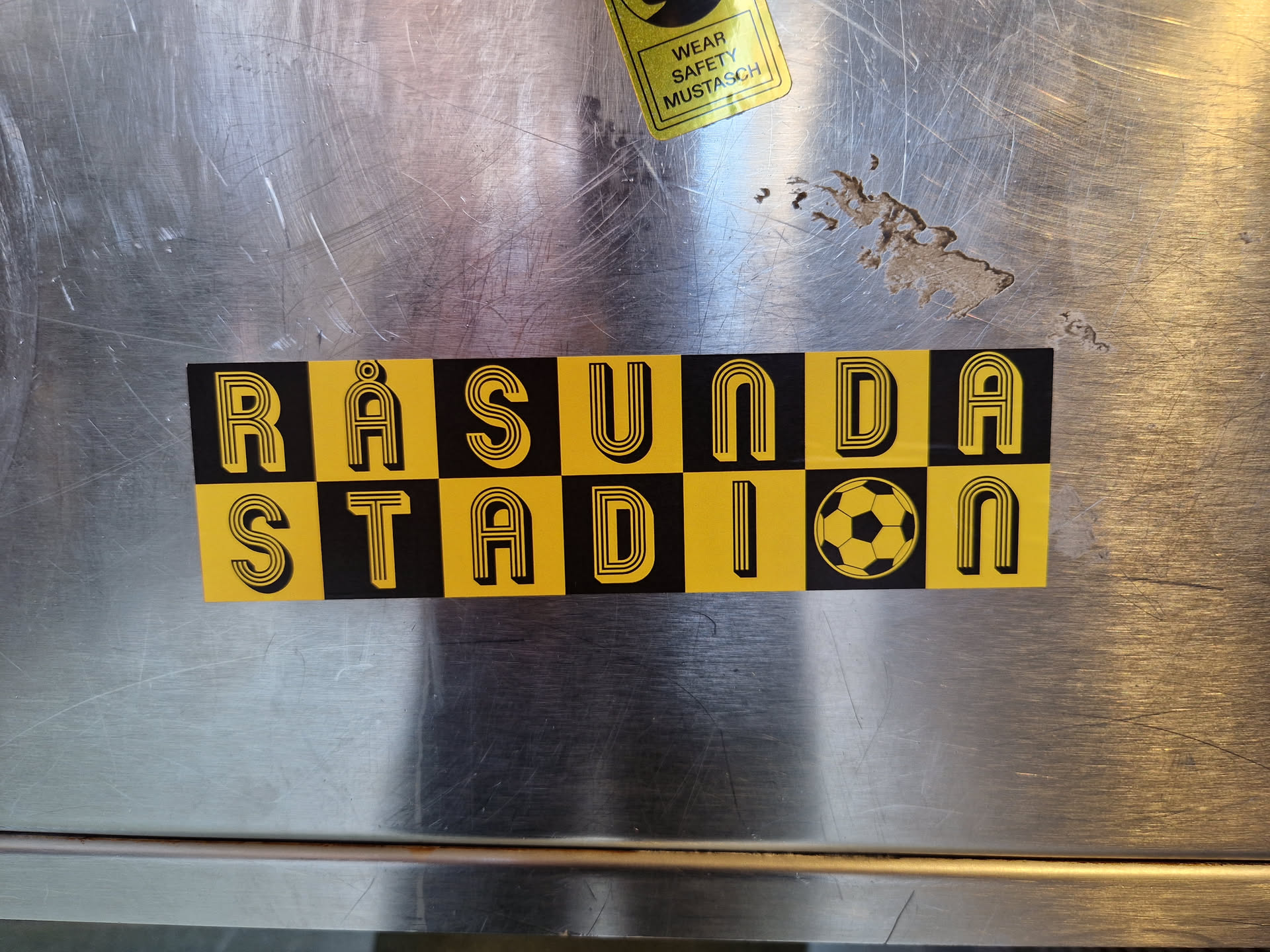 A yellow and black sticker with the words Räsunda Stadion and a soccer ball in the center. The text is in a retro style font.