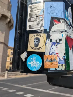 A close-up of a black pole covered with various stickers. The most prominent stickers include a circular sticker with a portrait and the text "MAZON CRIMINAL", and a round blue sticker with the word "Fresco" in white script. Other stickers include abstract designs and symbols, creating a dense and layered effect.