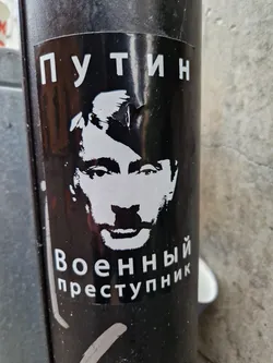 Black and white sticker featuring a stylized portrait of Vladimir Putin with the text "Putin" at the top and "War Criminal" at the bottom in Russian (Путин Военный преступник).