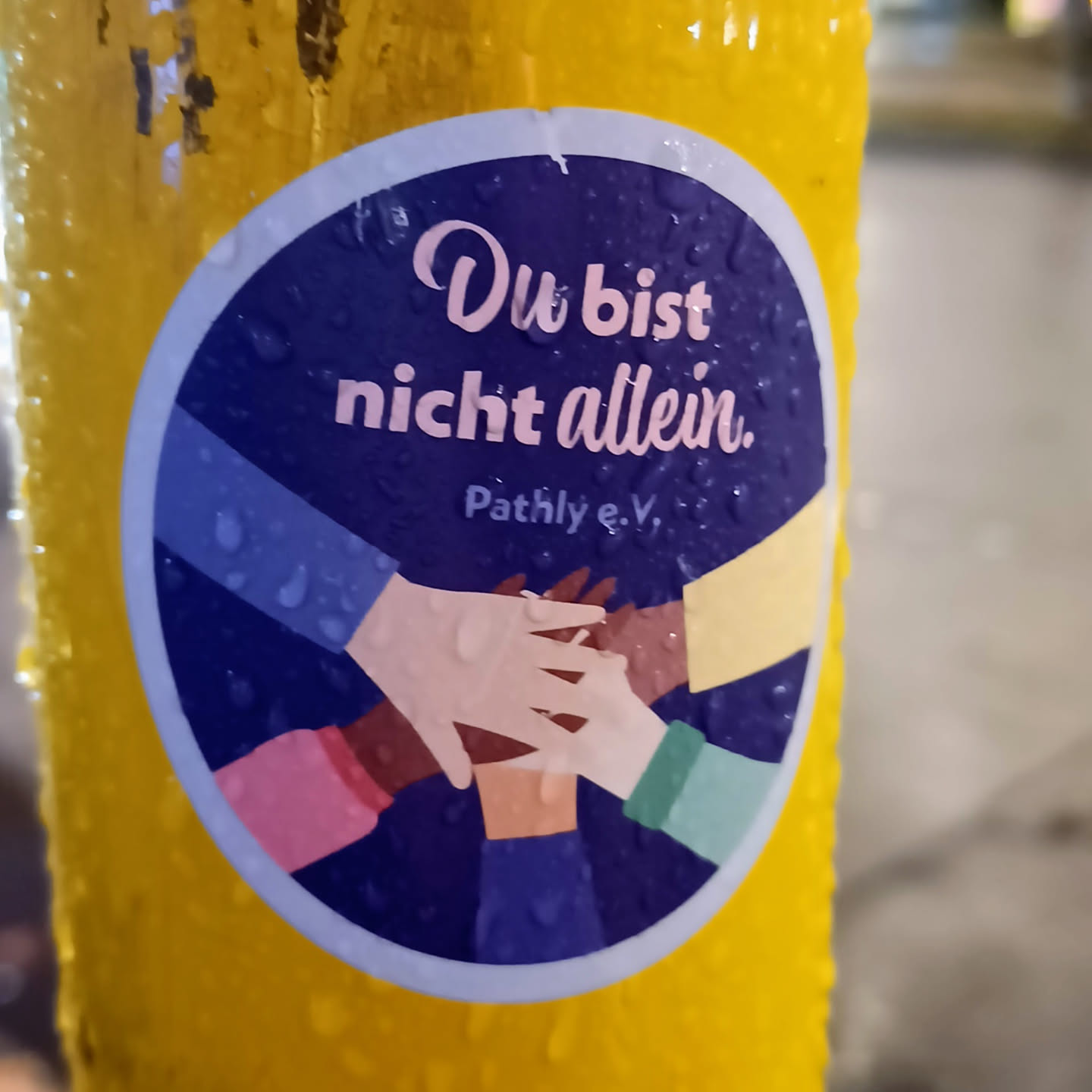 A round sticker on a yellow surface with a dark blue background.  The sticker shows several hands of different skin tones stacked on top of each other. The text 'Du bist nicht allein' (You are not alone) is visible in pink letters. 'Pathly e.V.' is written in a smaller font below the main text.