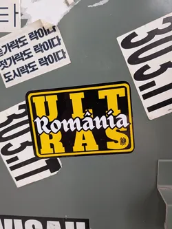 A rectangular sticker with rounded corners featuring a black background bordered by a thin yellow frame. The word 'ULTRAS' is displayed in bold, yellow block letters, split between the top row ('ULT') and the bottom row ('RAS'). Superimposed across the center is the word 'România' in a stylized white Fraktur-inspired Gothic font with black outlines. A small monogram icon appears inside the bottom right letter 'S'.