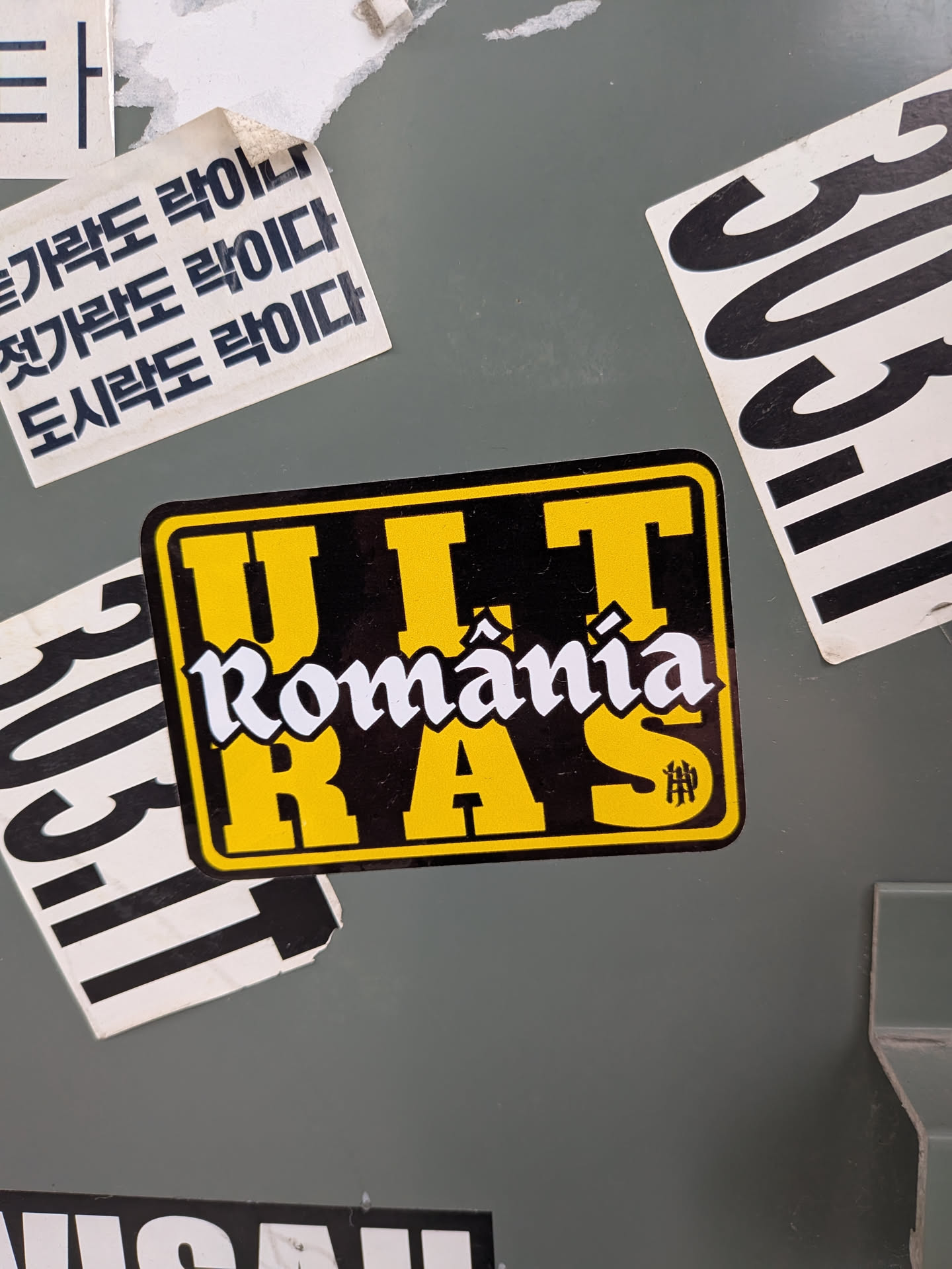 A rectangular sticker with rounded corners featuring a black background bordered by a thin yellow frame. The word 'ULTRAS' is displayed in bold, yellow block letters, split between the top row ('ULT') and the bottom row ('RAS'). Superimposed across the center is the word 'România' in a stylized white Fraktur-inspired Gothic font with black outlines. A small monogram icon appears inside the bottom right letter 'S'.
