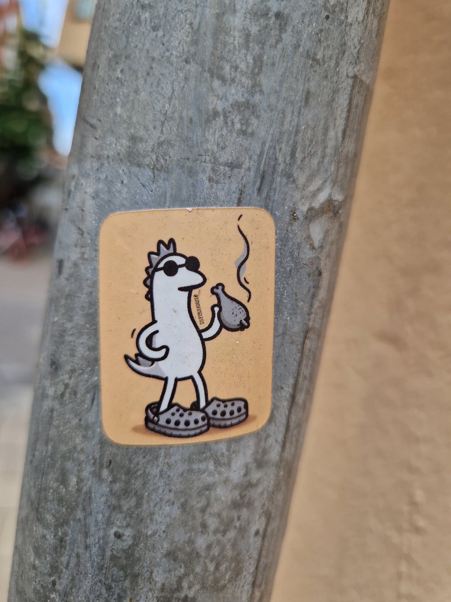 A sticker featuring a cartoon dinosaur wearing sunglasses and Crocs, smoking a pipe.  The dinosaur is white with a small crown, and the background is a light orange.