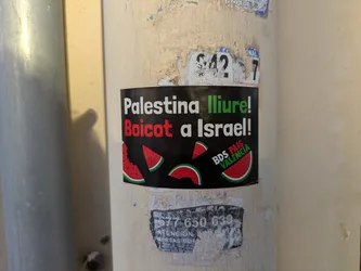 The sticker features the text "Palestina lliure! Boicot a Israel!" in black and red on a black background. Watermelon slices are placed strategically around the text. The text "BDS PAÍS VALENCIA" appears on the sticker. The sticker is attached to a light beige pole. Another partially destroyed sticker is below it, showing "577 650 639 ATENCION PUERTAS MOT".