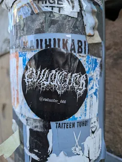 A circular, matte black sticker featuring a highly stylized, jagged white text in the center that reads 'EVILUCIFER'. Below this main logo, in a smaller, more legible but still gothic-style white font, is the social media handle '@evilucifer_666'. The sticker has a stark monochrome design, contrasting sharply with the layered, colorful, and worn stickers and posters on the pole it's adhered to.