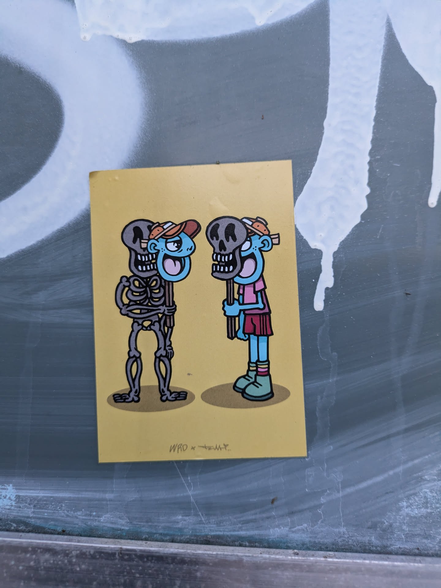 A yellow sticker featuring a whimsical illustration of two light-blue figures. One is a skeleton, the other appears human-like. Both hold a skull. The artist's signature or tag, 'WRO x TROLLY', is visible at the bottom.