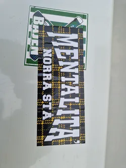 A black and yellow plaid sticker with the word "MENTALITA" written in white bold letters. There is a smaller sticker above it, partially visible, showing green and white colors.  The main sticker has a slightly worn look, suggesting it may have been applied outdoors for a while.